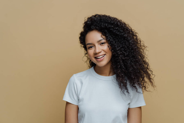 Portrait of beautiful woman with frizzy bushy hair, gentle smile and healthy skin, wears casual white t shirt, enjoys pleasant conversation stands against beige background copy space. People, emotions - 写真・画像