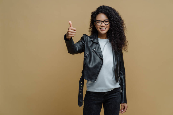 Zelfverzekerde vrolijke Afro-Amerikaanse vrouw toont duim omhoog in goedkeuring, geeft aanbeveling of advies gekleed in stijlvolle leren jas glimlacht positief geïsoleerd op beige achtergrond. Ik hou van en accepteer - Foto, afbeelding