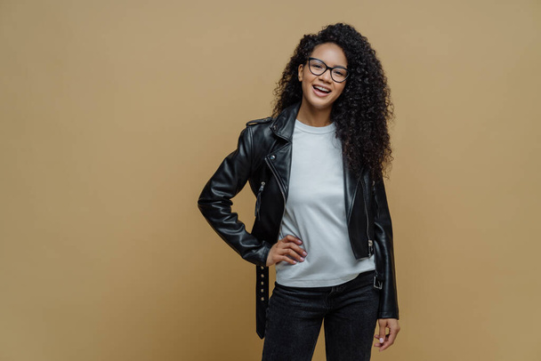 Fashionable woman with glad expression, keeps hand on hip, smiles cheerfully, wears optical glasses, white t shirt, black leather jacket and jeans, ready for walk, poses against beige background - Photo, Image