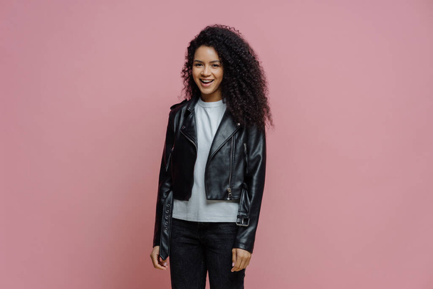 Happy positive Afro American woman dressed in leather jacket, ready for outdoor walk, poses against pink background. Joyful dark skinned millennial girl in good mood has casual talk with friend - Φωτογραφία, εικόνα