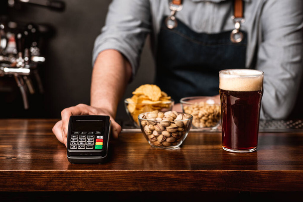 Modern payment at bar. Barman in apron gives to customer a glass of dark beer, snacks and terminal - Photo, Image