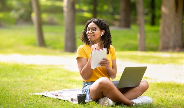 Positives schwarzes Mädchen mit Laptop lernt online im grünen Sommerpark, leerer Raum - Foto, Bild