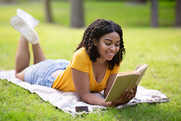 Χαρούμενη Αφροαμερικανή γυναίκα διαβάζει ενδιαφέρον βιβλίο, ενώ βρίσκεται στο πράσινο γκαζόν στο πάρκο - Φωτογραφία, εικόνα