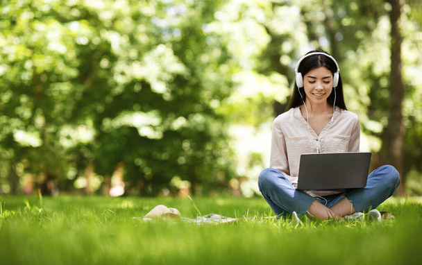 Gioioso asiatico ragazza seduta su prato con computer portatile, studiare e ascoltare musica - Foto, immagini