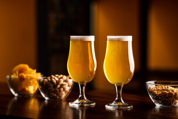 Два бокала с легким пивом и пеной на деревянном барной стойке в интерьере паба, фисташки, крекеры и орехи в стеклянных тарелках поблизости
 - Фото, изображение