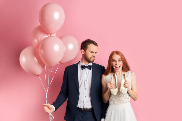 Unpleased man in tuxedo hold pink air balloons while woman hold shoes - Zdjęcie, obraz