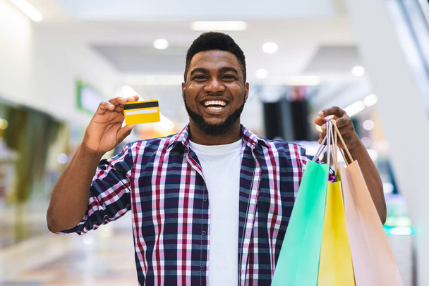 Pago fácil. Hombre negro mostrando tarjetas de crédito y bolsas de compras en el centro comercial
 - Foto, Imagen