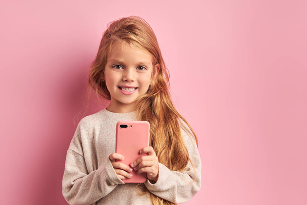 Smiling sweet little girl with long hair use smartphone on pink background - Foto, imagen