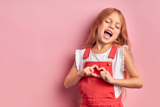 Funny little girl show heart isolated over pink background - Zdjęcie, obraz