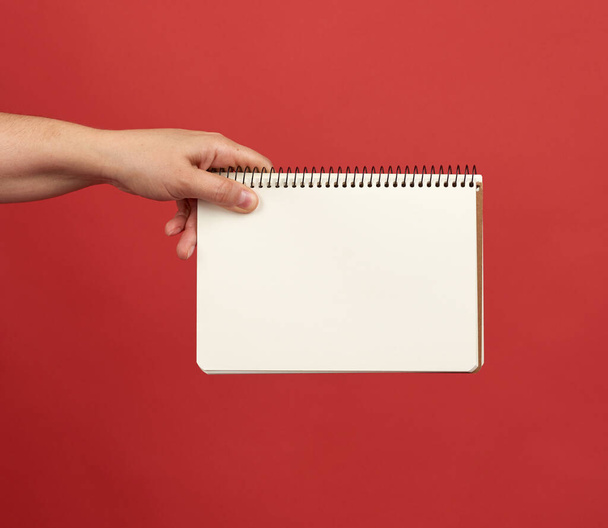 main tenant un carnet ouvert en spirale avec feuilles blanches vides, fond rouge, espace de copie - Photo, image