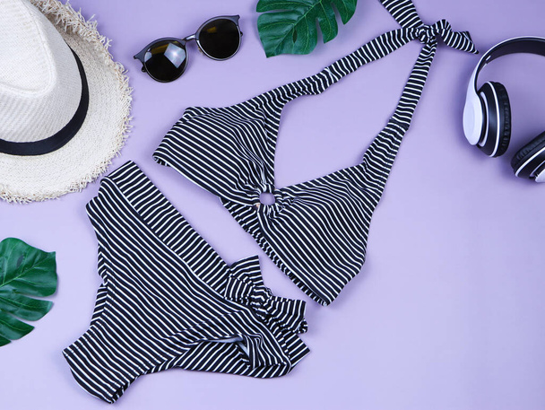 Utazási és nyári nyaralás koncepció, felső nézet fekete-fehér csíkos minta bikini fürdőruha és a női nyaralás tartozékok tételek lila háttér trópusi szörnylevelek. - Fotó, kép