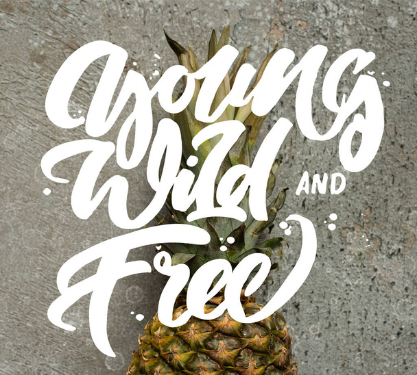top view of ripe pineapple on grey concrete surface with young, wild and free illustration - Foto, imagen