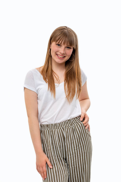 Half length portrait of a young girl with long blonde hair with a bang wearing  white shirt and striped pants against white background in studio - Foto, Imagem