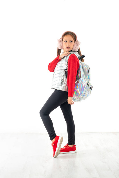 Ganzkörperporträt eines jungen ernsten Mädchens, das eine glänzende Jacke trägt und mit einem Rucksack ähnlicher Farbe unterwegs ist, isoliert.  - Foto, Bild