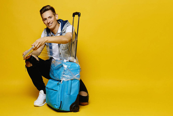 A young man in denim clothes is preparing his travel suitcase to give it into the luggage, isolated at the yellow background. Mockup of travel advertise - Fotografie, Obrázek
