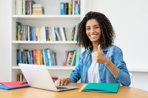 Latin female student with curly hair at computer showing thumb up at desk at home - Fotó, kép