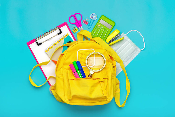 Powrót do szkoły, koncepcja edukacji Żółty plecak ze sprzętem szkolnym, ochronna maska medyczna, kalkulator, nożyczki odizolowane na niebieskim tle. Widok z góry Kopiuj przestrzeń Flat lay - Zdjęcie, obraz