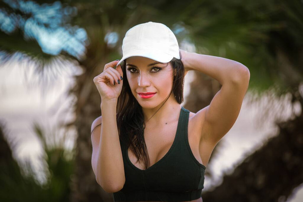 Plajda palmiye ağaçlarıyla çevrili beyaz şapkalı genç ve çekici bir kadın. - Fotoğraf, Görsel