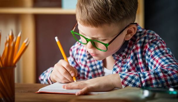 Χαρούμενο χαμογελαστό αγόρι με γυαλιά στο σχολείο για πρώτη φορά. Παιδί σε εσωτερικούς χώρους στο σπίτι στο εσωτερικό του δωματίου του παιδιού με ξύλινο υπόβαθρο. Πίσω στο σχολείο. Το αγοράκι γράφει στο κενό. Εκπαιδευτική έννοια. - Φωτογραφία, εικόνα