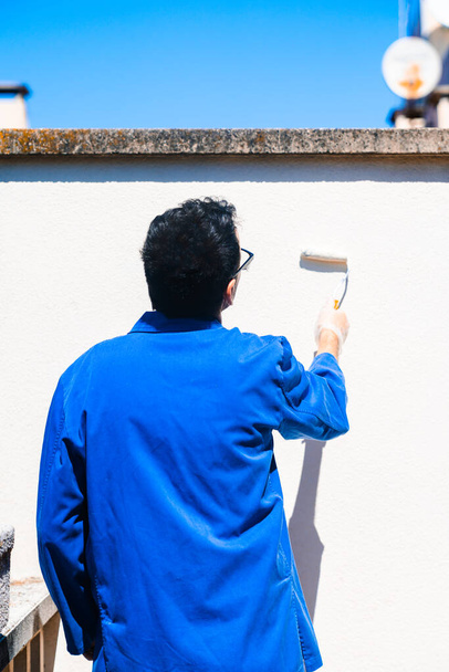 Painter working on painting the white wall of the house terrace. Back view of mature man reforming the home. - Photo, Image