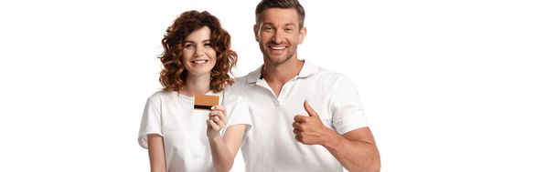 白で隔絶されたクレジットカードを持っている妻の近くに親指を示す幸せな男のパノラマショット  - 写真・画像