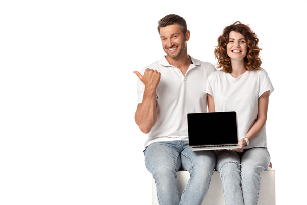 cheerful woman holding laptop with blank screen while man pointing with thumb isolated on white  - Foto, Imagem