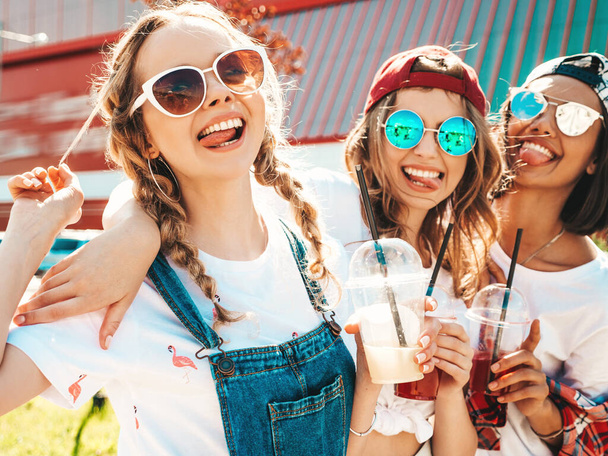 Three young beautiful smiling hipster girls in trendy summer clothes.Sexy carefree women posing outdoors.Positive models holding and drinking fresh cocktail smoothie drink in plastic cup with straw - Foto, Imagem