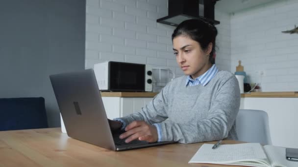 Nuori intialainen nainen työskentelee kotona, toimii tietokoneella ja kirjoittaa muistiinpanoja muistikirjaan. Hänellä on paita ja villapaita. - Materiaali, video