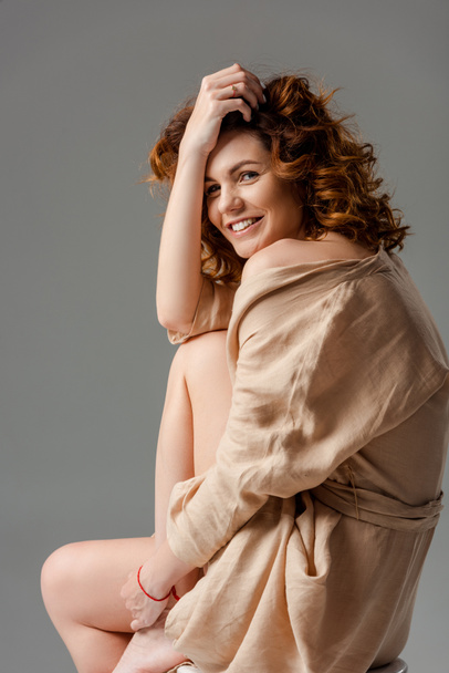 curly woman smiling while sitting on chair isolated on grey  - Photo, Image