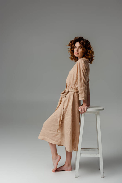 curly and barefoot woman standing near chair on grey  - Foto, imagen