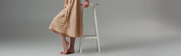 panoramic crop of barefoot woman standing near chair on grey  - Foto, Imagem