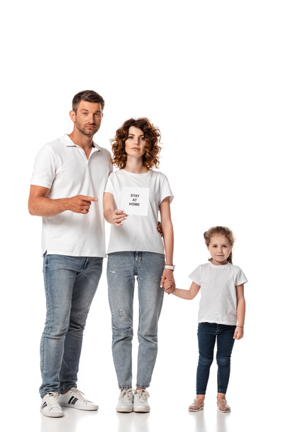 handsome man pointing with finger at card with stay at home lettering near attractive wife and kid on white  - Fotoğraf, Görsel