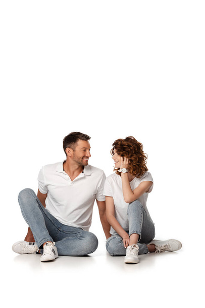 happy couple in denim jeans sitting and looking at each other on white - Фото, зображення