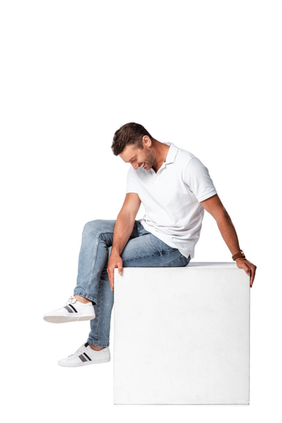 glücklicher Mann in Jeans, der auf Würfel sitzt und isoliert auf Weiß herabblickt - Foto, Bild