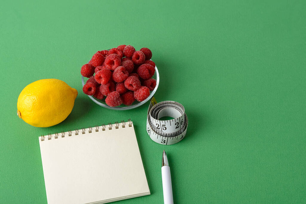 Inch measuring tape roll, lemon, raspberries, paper notepad and white pen on a green background. Concept of healthy eating, diet, weight loss and body slim. Copy space.  - Foto, imagen
