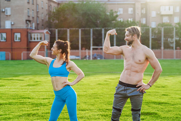 Exercises in pairs. Attractive athletes girl and guy demonstrating pumped up body. Guy is standing with a naked torso, girl is wearing a blue top and leggings. Showing muscles on the arms. - Fotó, kép