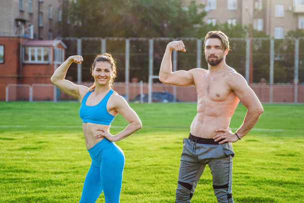 Exercises in pairs. Attractive athletes girl and guy demonstrating pumped up body. Guy is standing with a naked torso, girl is wearing a blue top and leggings. Showing muscles on the arms. - Фото, изображение