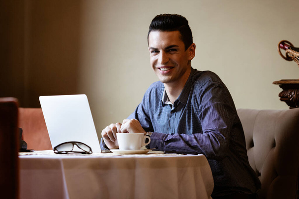 LGBTQ community lifestyle concept. Young homosexual man sits at the table in old-fashioned city cafe. Handsome gay male businessman works on laptop computer while having a break at lunch. - Фото, изображение