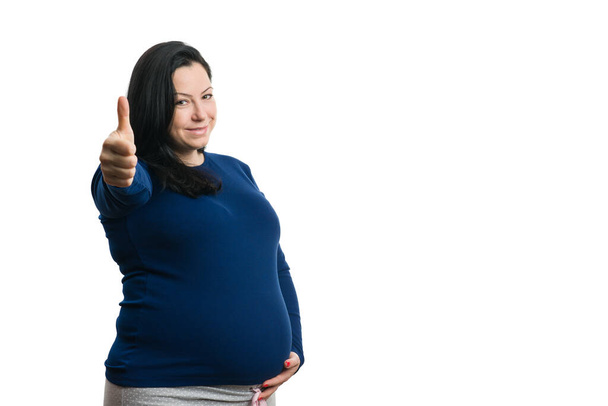 Mutlu ifadeli bir kadın, beyaz arka planda izole edilmiş hamile göbeğine dokunarak el hareketi yapıyor. - Fotoğraf, Görsel