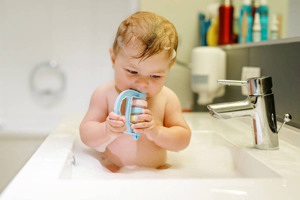 Cute adorable baby taking bath in washing sink and grab water tap. Little healthy girl with big blue eyes having fun and playing with soap foam. - Photo, Image