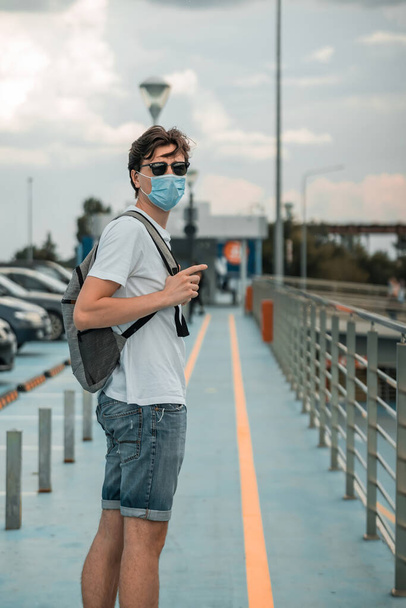 Young man in white t-shirt, jeans shorts, sunglasses, surgical mask and backpack on his back looking back in the parking lot of the airport. Travelling during coronavirus pandemy outbreak - Foto, afbeelding