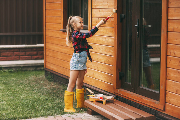 Menina adolescente bonita em macacão jeans e uma camisa xadrez está pintando sua casa de madeira. Melhoria da casa, reparação de DIY
 - Foto, Imagem