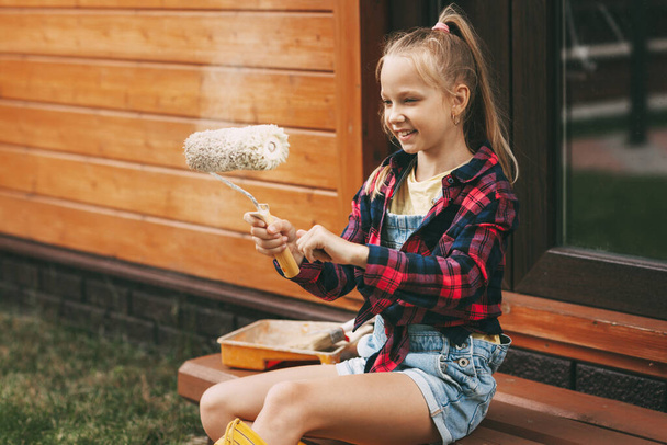 Beautiful teenage girl in denim overalls and a plaid shirt is roller-painting her wooden house. Home Improvement, DIY Repair - Photo, image
