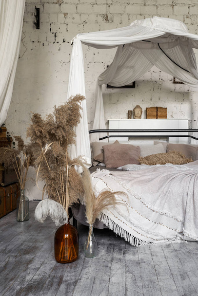 Yatağın üzerinde tepe örtüsü, ahşap zeminde vazodaki kuru bitkiler ve ev dekoruyla bohem tarzında rahat beyaz yatak odasının dikey fotoğrafı. - Fotoğraf, Görsel