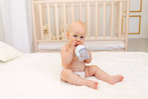 baby boy 8 months old sitting in diapers on a white bed with a bottle of milk at home, baby food concept, first bait, place for text - 写真・画像