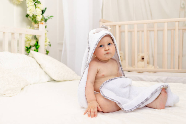 chłopiec 8 miesięcy siedzi na białym łóżku w białym ręczniku po kąpieli w łazience w domu, miejsce na tekst - Zdjęcie, obraz