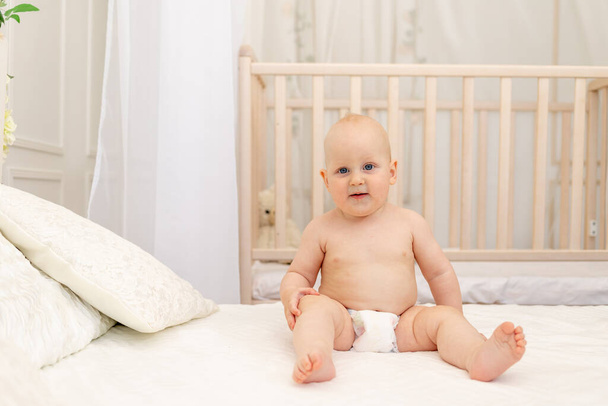 hymyilevä poikavauva 8 kuukauden ikäinen istuu vaipoissa valkoisella sängyllä, jossa on pullo maitoa kotona, vauvanruokakonsepti, ensimmäinen syötti, tekstipaikka - Valokuva, kuva