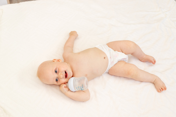 bebé niño de 8 meses de edad acostado en pañales en una cama blanca con una botella de leche en casa piernas arriba, vista superior, concepto de alimentos para bebés, bebé bebiendo agua de un biberón, lugar para texto - Foto, Imagen