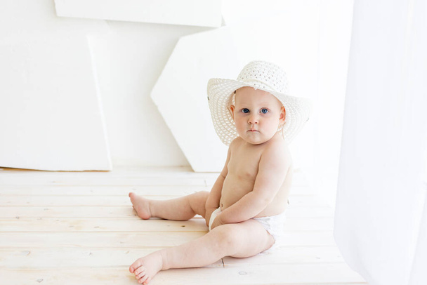niño de 8 meses sentado en pañales con un sombrero blanco en la ventana, lugar para el texto
 - Foto, imagen