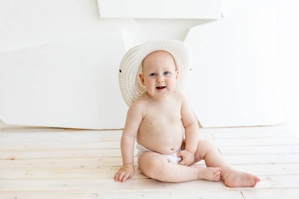8ヶ月前にオムツを着た赤ちゃんが窓の白い帽子に座っていました - 写真・画像